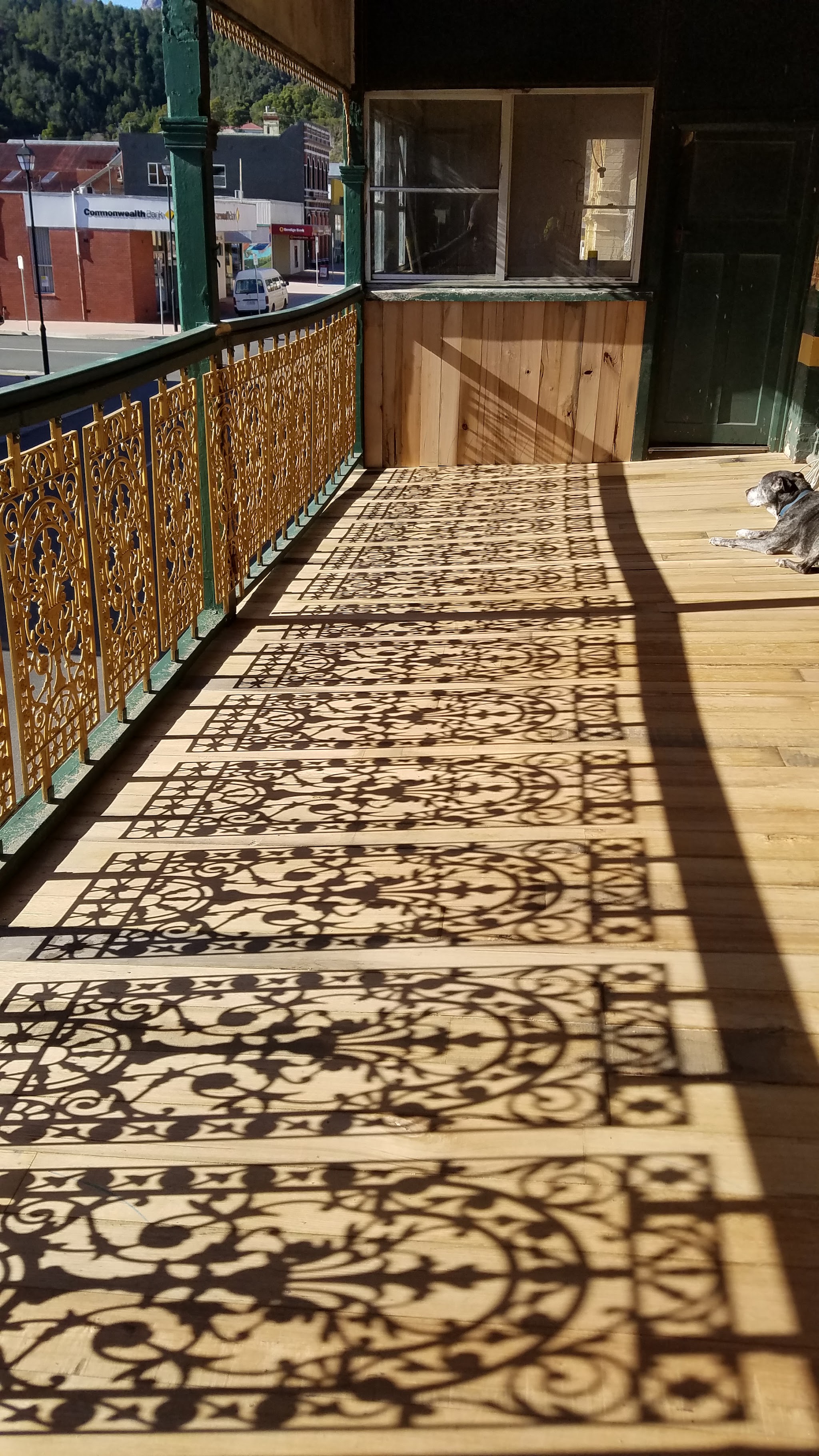 Gold cast iron lacework along the hotel balcony, casting patterned shadows onto new timber decking, with a dog asleep in the sun
